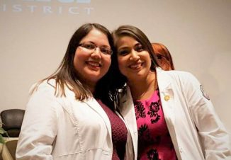 Graduates Juana and Araceli hug and smile at their pinning ceremony in their nurse lab coats.