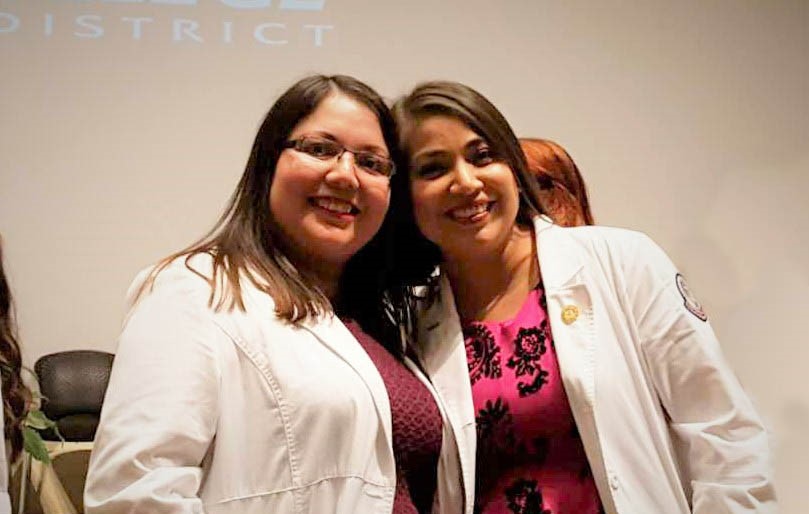 Graduates Juana and Araceli hug and smile at their pinning ceremony in their nurse lab coats.