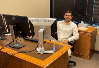 Graduate Khaibar Rahimy sits at his desk at work. He smiles behind three large computer monitors.