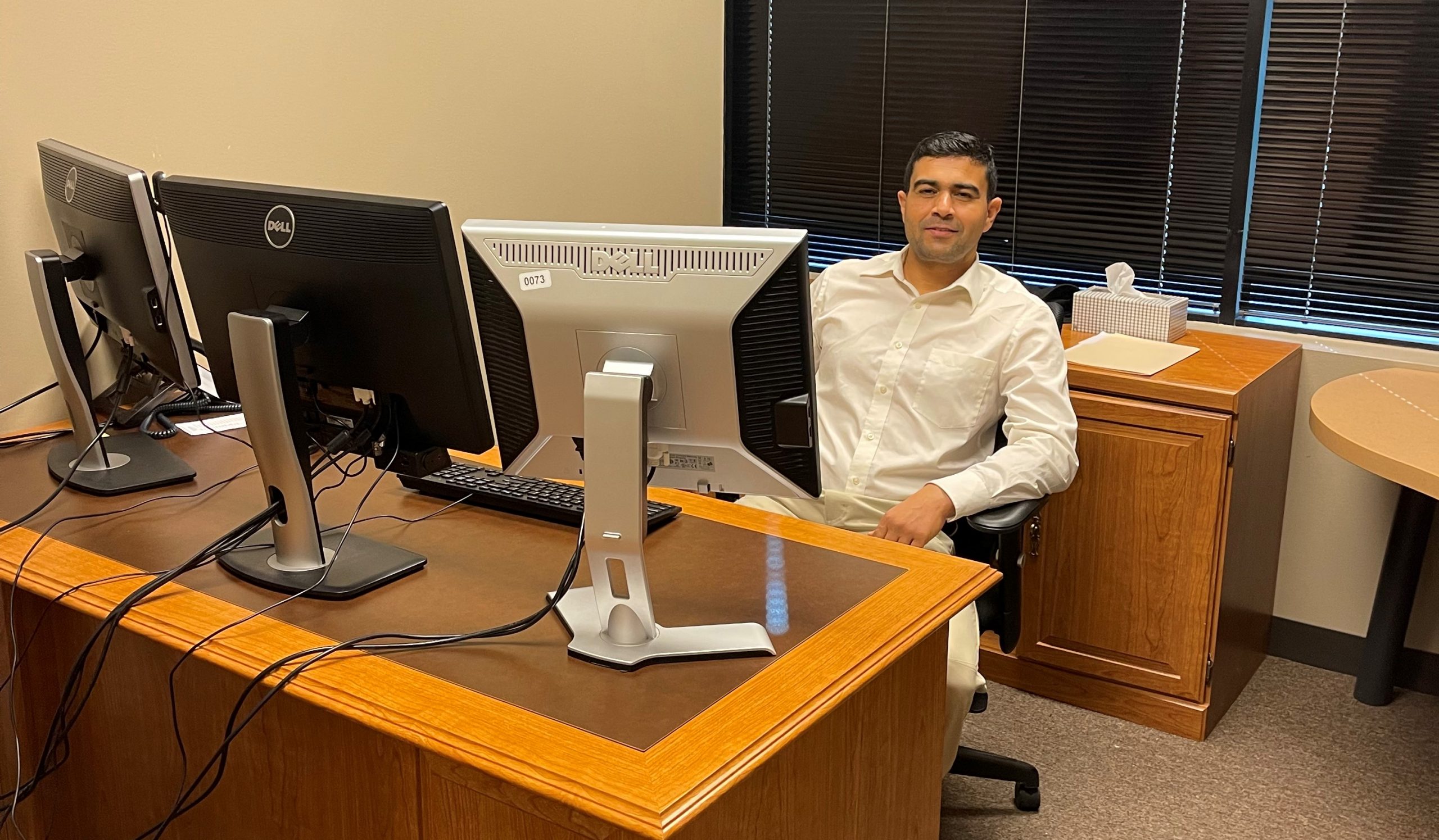 Graduate Khaibar Rahimy sits at his desk at work. He smiles behind three large computer monitors.
